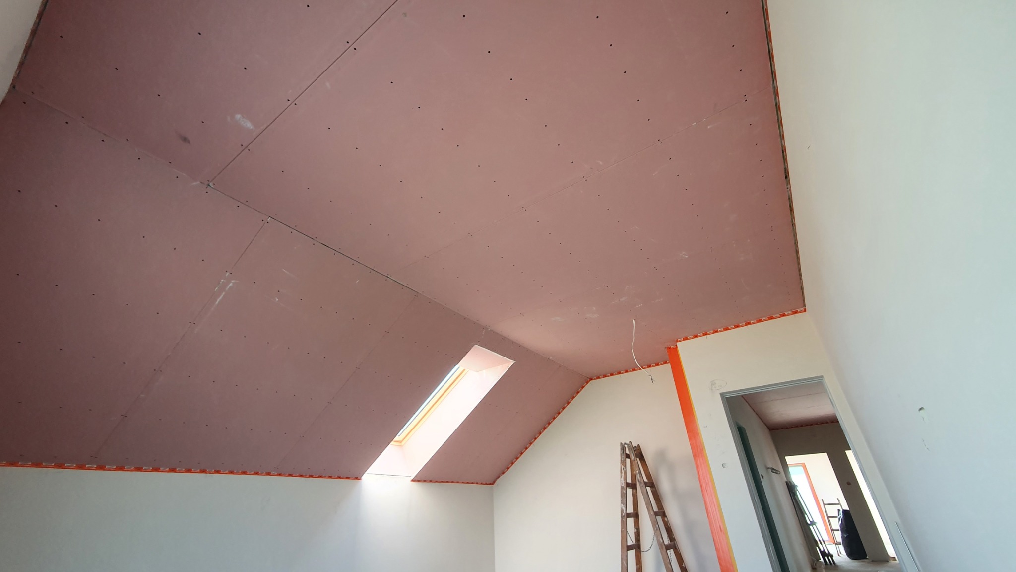 Montaż płyt karton-gips na poddaszu ze świetlikiem. Widoczne łączenia płyt i taśma zabezpieczająca. Drabina oparta o ścianę. Surowy stan wykończenia wnętrza.