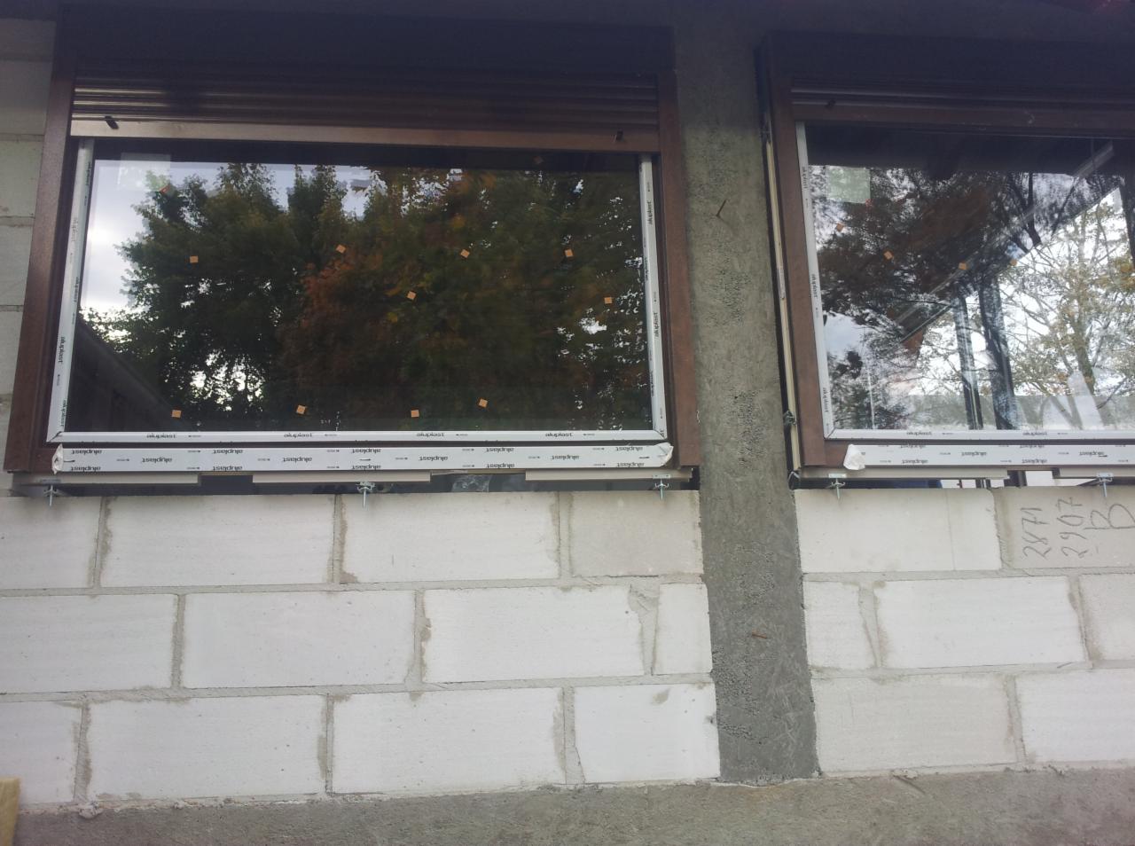 Montaż okien z roletami zewnętrznymi w surowym budynku z betonu komórkowego. Widoczne śruby mocujące i folia ochronna na profilach.