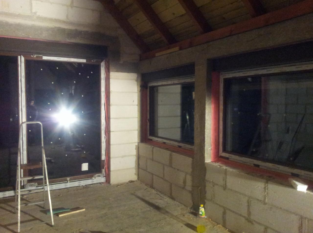 Montaż okien i bramy garażowej w surowym garażu z bloków. Widoczna drabina i częściowo wykonane obramowanie. Oświetlenie odbija się w szybach.