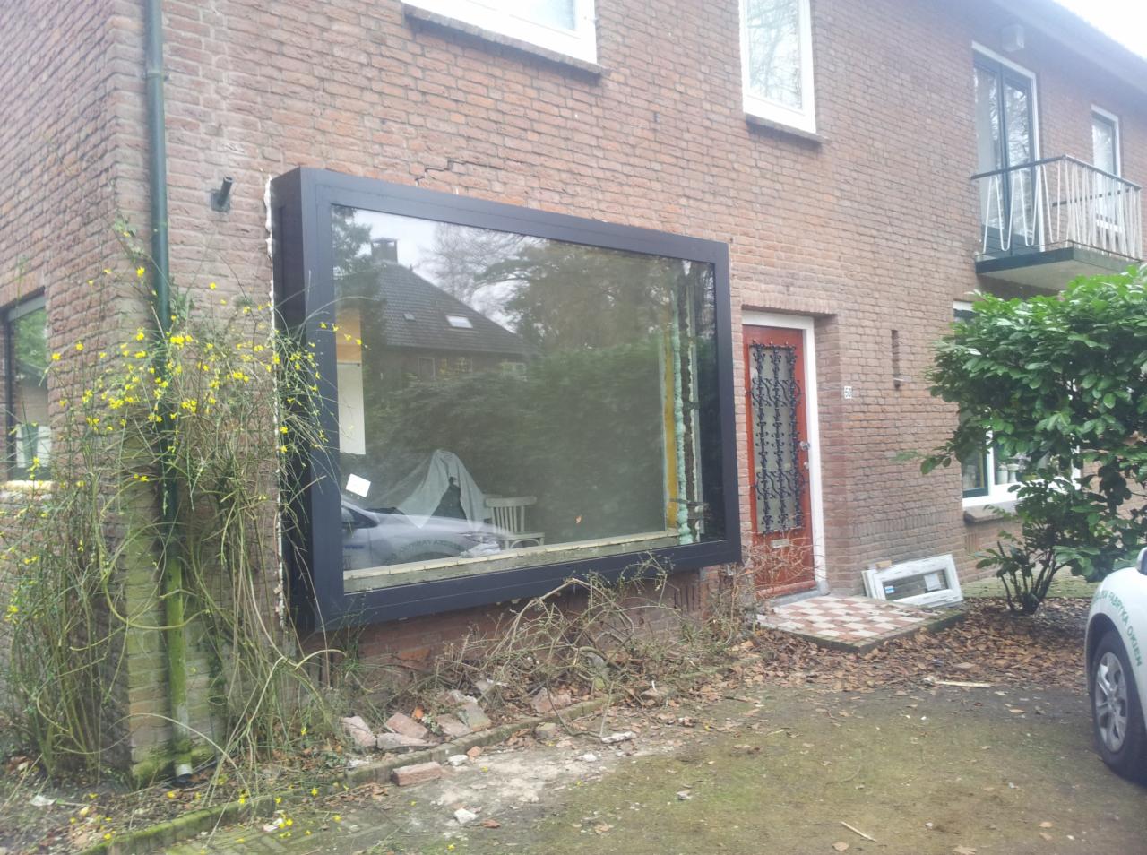 Ceglany budynek z dużym oknem w czarnej ramie, odbijającym krajobraz. Widoczne zarośla i fragment samochodu. Elementy demontażu przy wejściu.