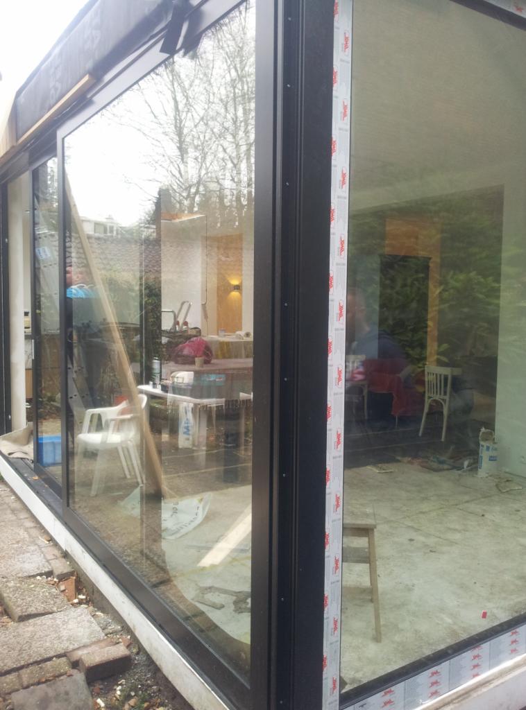 Montaż okna tarasowego z czarną ramą. Widoczne akcesoria montażowe i wnętrze domu w odbiciu szyby. Okno z folią ochronną.