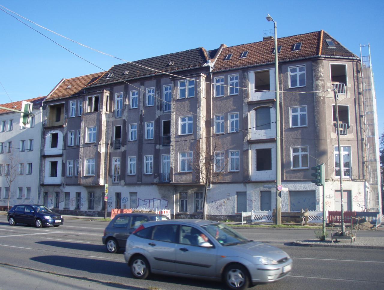 Kamienica w trakcie remontu z odsłoniętymi otworami okiennymi, widoczna z ulicy z przejeżdżającymi samochodami w słoneczny dzień.