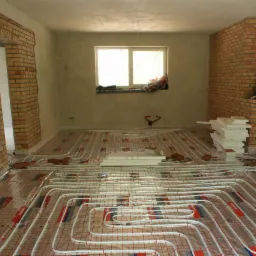 Instalacja ogrzewania podłogowego w Koluszkach. Białe rury ułożone na folii z czerwoną siatką, ceglane ściany w tle. Etap prac przed wylewką.