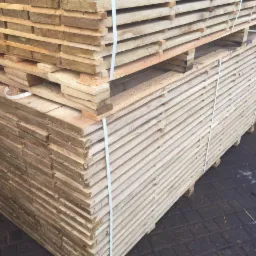 Stos desek budowlanych na paletach, zabezpieczony białymi taśmami. Widoczne słoje drewna i tekstura. Tło stanowią deski i szara kostka brukowa.