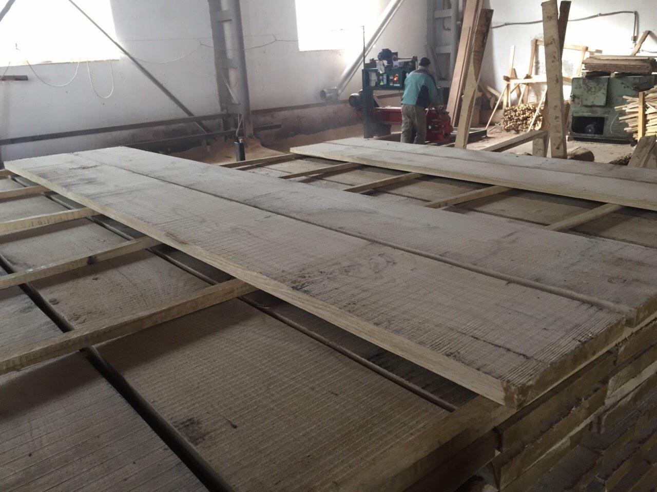Stos desek w magazynie drewna, w tle pracownicy obsługujący maszynę do obróbki drewna. Widoczne ślady obróbki na deskach, surowe drewno.
