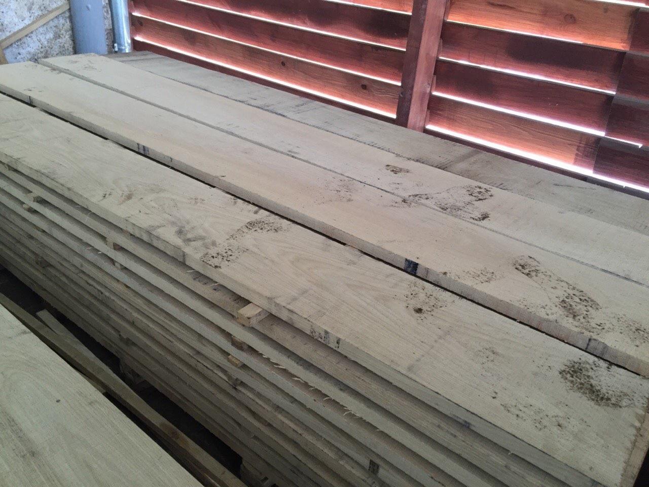 Stos desek z jasnego drewna, widoczne ślady po wodzie, ułożone równo na tle drewnianej ściany z poziomymi szczelinami przepuszczającymi światło.