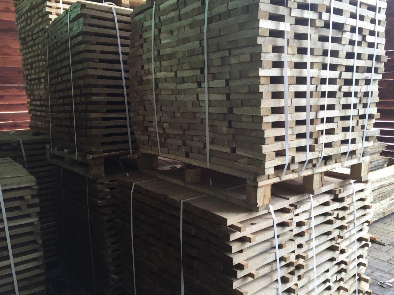 Stosy jasnych desek ułożonych na paletach, zabezpieczonych białymi taśmami, ukazane w magazynie drewna. Widoczne struktury drewna i regularny układ desek.