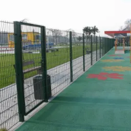 Zielone ogrodzenie panelowe z furtką oddziela plac zabaw z kolorowymi kwiatami od parku. Widoczna ławka i kosz na śmieci za ogrodzeniem.