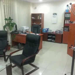 Jasne pomieszczenie biurowe z biurkiem, krzesłami, szafkami i wieszakiem na ubrania, roślina doniczkowa przy oknie.