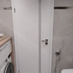 Białe skrzydło drzwiowe z przeszkleniem w łazience, widoczne elementy armatury i pralka.