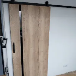 Drewniane drzwi przesuwne z czarnym uchwytem i systemem jezdnym zamontowane na białej ścianie, widoczny fragment podłogi i gniazdka elektrycznego.