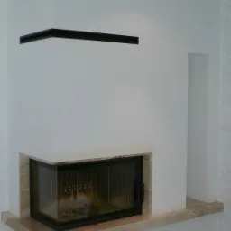 Narożny kominek z czarnymi akcentami, obudowany beżowym kamieniem. Minimalistyczny design, białe ściany. Widoczne palenisko i fragment podłogi.