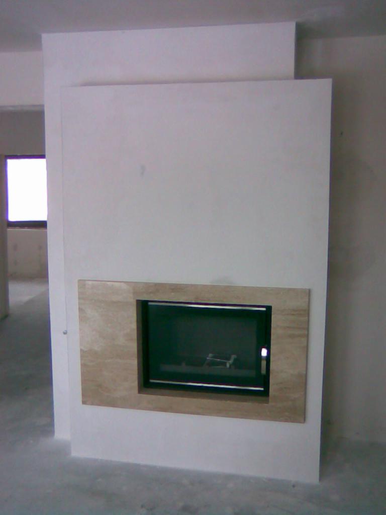 Wnękowy kominek z ramą w kolorze beżowym, osadzony w surowej, białej ścianie. Widoczny fragment pomieszczenia w stanie deweloperskim.