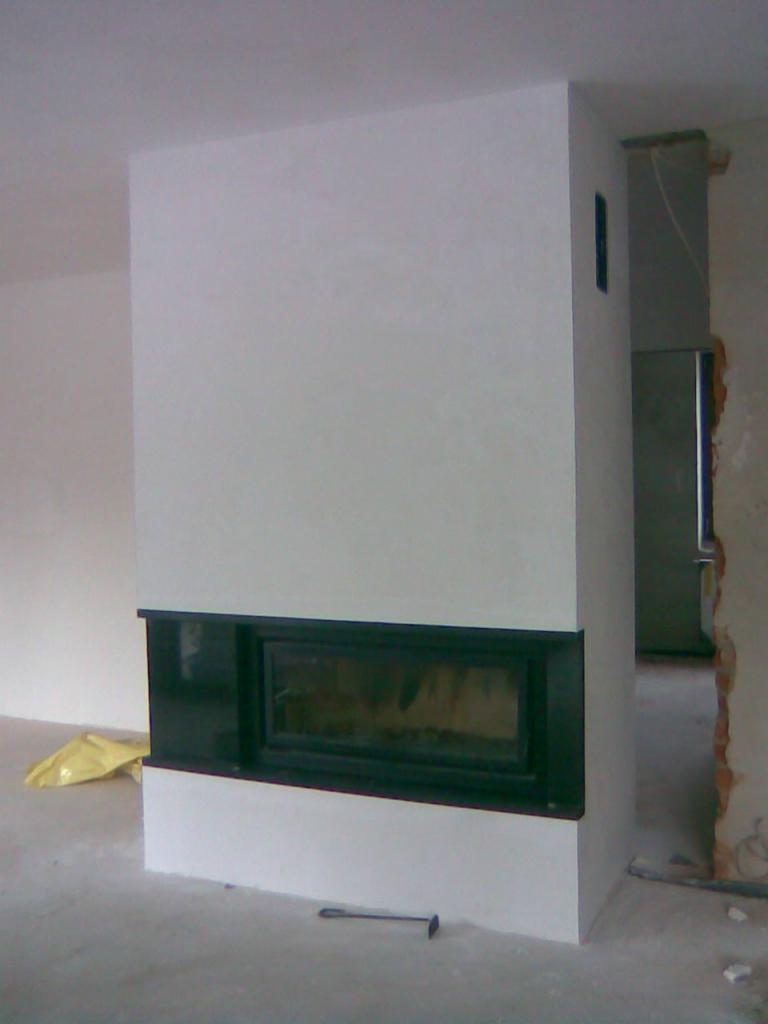 Nowoczesny kominek w trakcie montażu, czarna rama, biała obudowa, surowy stan wykończenia wnętrza, widoczne narzędzie na podłodze.