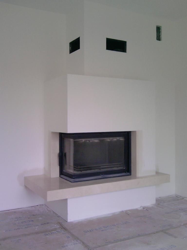 Nowoczesny kominek narożny z czarnym wkładem, obudowany jasnym kamieniem i płytami gipsowymi. Widoczne kratki wentylacyjne w ścianie nad kominkiem.