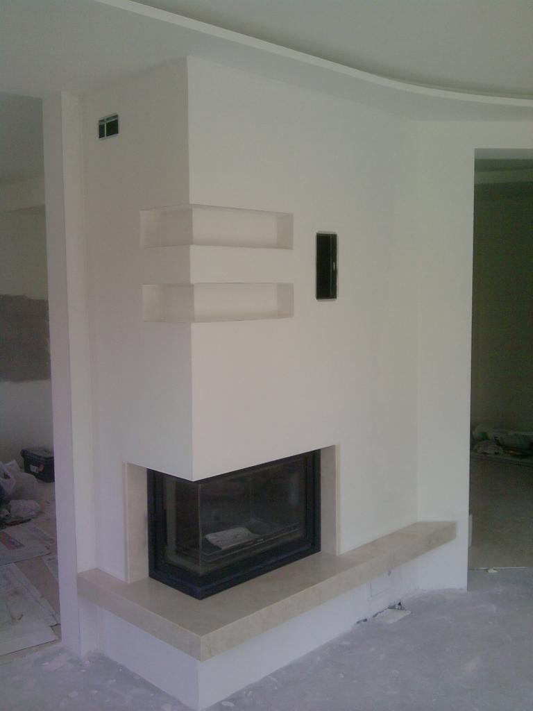 Nowoczesny kominek narożny z czarnym wkładem i jasnym kamieniem, wnęki dekoracyjne w ścianie, minimalistyczny design, białe ściany.