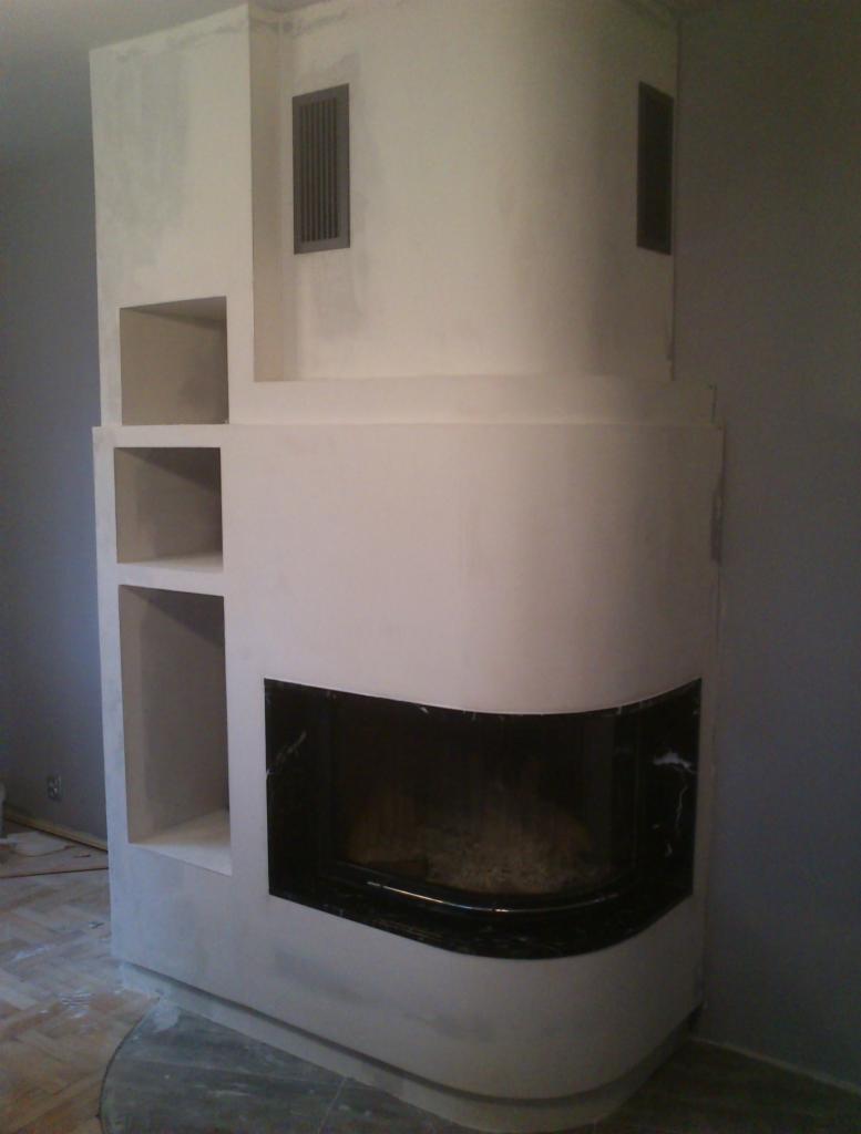 Narożny kominek w trakcie wykończenia, z półkami i czarnym obramowaniem paleniska. Widoczne detale wykończeniowe i fragment parkietu.