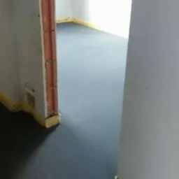 Gładka, szara posadzka w nowym pomieszczeniu z widocznymi elementami muru i taśmą zabezpieczającą przy ścianach, tworząca minimalistyczne wnętrze.