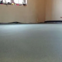 Świeżo wylana, jasnoszara posadzka w pomieszczeniu z oknem. Widoczne narzędzia przy ścianie i czarna folia zabezpieczająca na obrzeżach. Minimalistyczny styl.