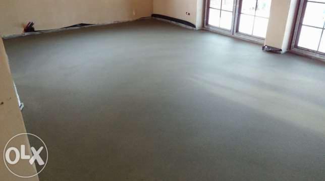 Świeżo wylana, gładka posadzka w jasnym pomieszczeniu z dużymi oknami. Widoczne kable przy ścianie, sugerujące prace instalacyjne przed wylaniem.