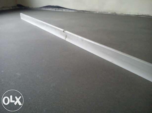 Świeżo wylana, szara posadzka, wyrównywana długą, aluminiową łatą. Widoczne ślady obróbki i gładka powierzchnia. W tle fragment ściany.