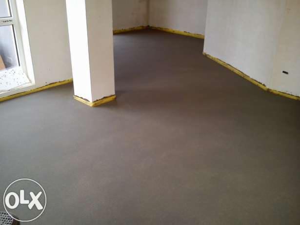 Świeżo wylana, gładka, szara posadzka w pomieszczeniu z białymi ścianami i żółtymi taśmami przy krawędziach. Widoczny biały filar w rogu pokoju.