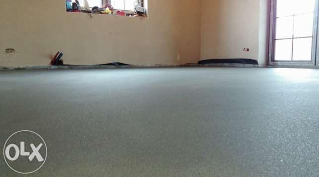 Świeżo wylana, jasnoszara posadzka w pomieszczeniu z oknem. Widoczne narzędzia przy ścianie i czarna folia zabezpieczająca na obrzeżach. Minimalistyczny styl.