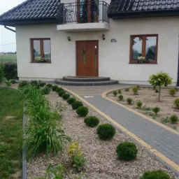 Dwukondygnacyjny dom z balkonem i brązowymi oknami, numer domu 67b, ścieżka z kostki brukowej prowadząca do wejścia, ozdobny ogród z kamykami i roślinami.