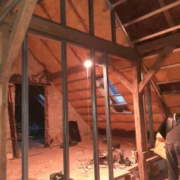 Konstrukcja metalowa szkieletu ścianki działowej na poddaszu z widocznym ociepleniem wełną mineralną, fragment muru z cegły, narzędzia i lampa oświetlająca przestrzeń.