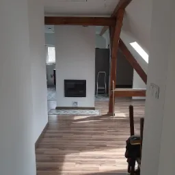 Wnętrze poddasza po remoncie z drewnianą konstrukcją stropu, widoczne podłogi z paneli, kominek i fragment kuchni w tle.