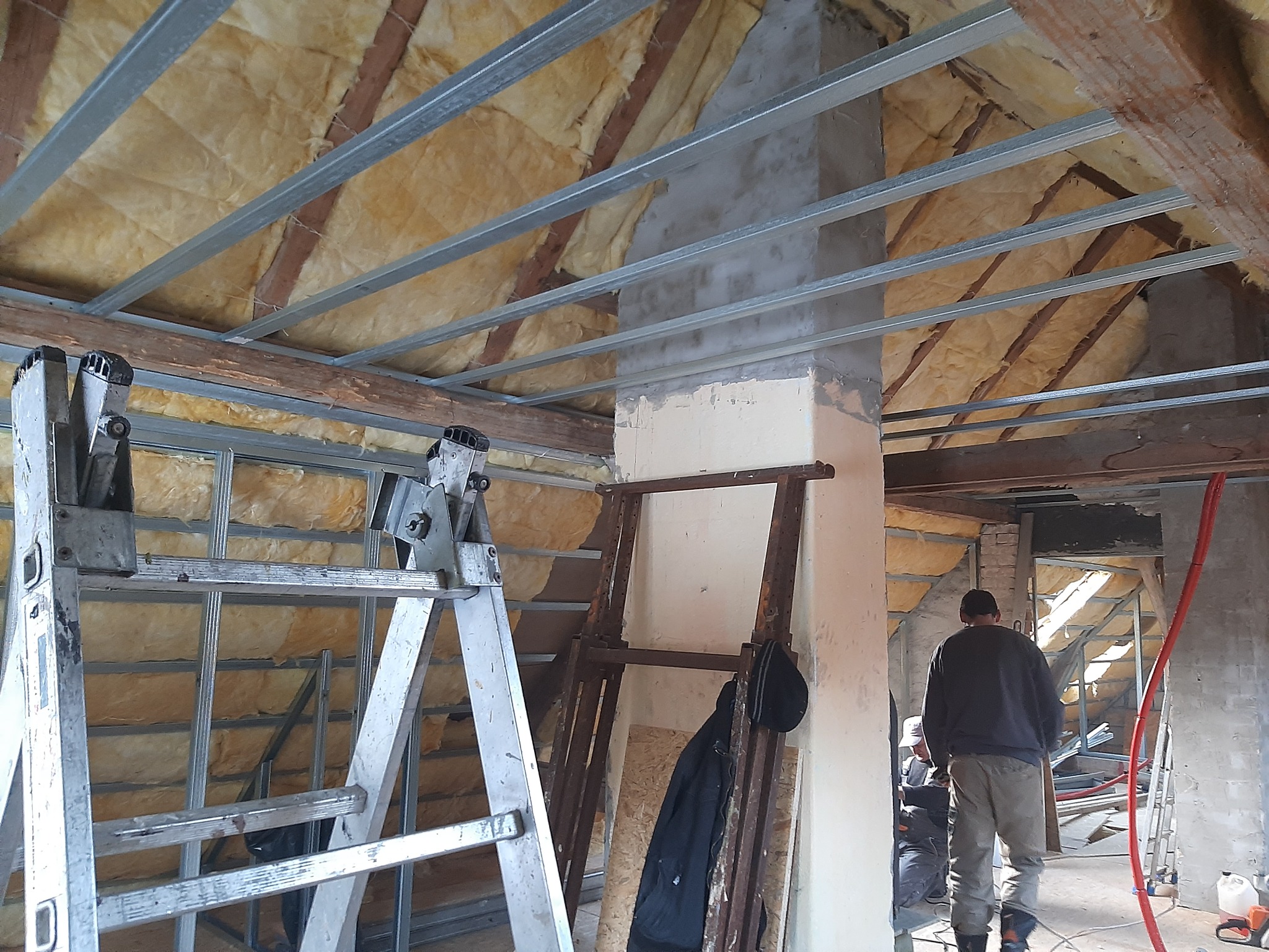 Ocieplenie poddasza wełną mineralną, widoczne profile metalowe, drabiny i pracownicy w tle, surowy stan wykończenia.