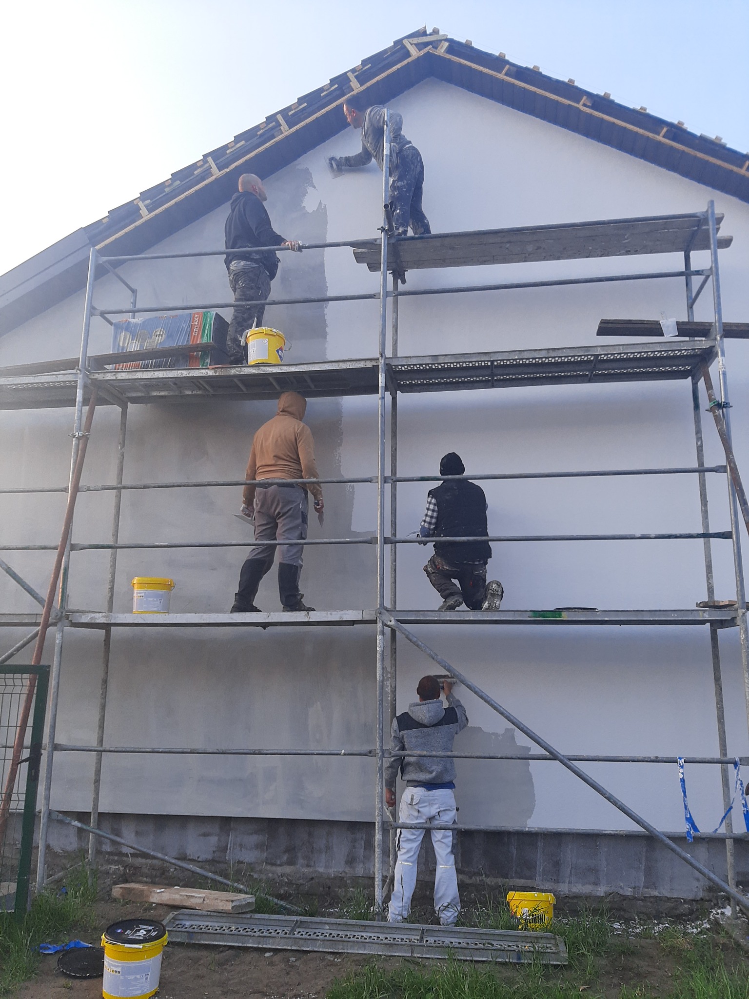 Ekipa tynkarzy na rusztowaniu podczas nakładania tynku na elewację domu jednorodzinnego, widoczne żółte wiadra z tynkiem i narzędzia.