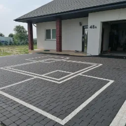 Ułożona, grafitowa kostka brukowa z białymi liniami tworzącymi geometryczny wzór przed domem z numerem 48A i otwartym garażem po prawej stronie.