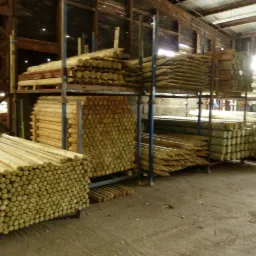 Magazyn z ułożonymi na metalowych regałach okrąglakami i kantówką, niektóre okrąglaki zaostrzone na końcach.