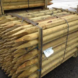 Stos impregnowanych, zaostrzonych drewnianych słupk&oacute;w ogrodzeniowych, zabezpieczonych taśmami, z widoczną etykietą producenta.