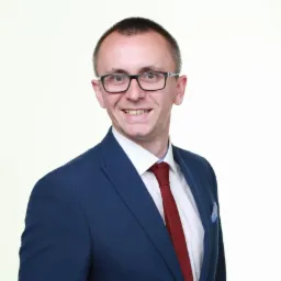 Uśmiechnięty mężczyzna w okularach, granatowej marynarce i czerwonym krawacie, portret biznesowy na jasnym tle. Widoczna poszetka w kieszonce marynarki.