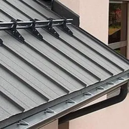 Szara blacha na dachu domu jednorodzinnego z czarnymi rynnami i uchwytami przeciwśniegowymi. Widok z boku, z fragmentem elewacji i otoczenia.