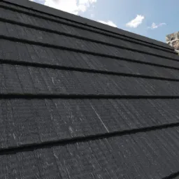 Czarny dach z dachówkami o strukturze drewna, widoczny komin z kamienia na tle błękitnego nieba z chmurami. Ujęcie pod kątem, eksponuje fakturę pokrycia.