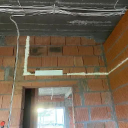 Instalacja elektryczna w budynku w trakcie budowy; widoczne przewody na suficie, puszka elektryczna zamontowana w ścianie z cegły oraz pianka montażowa uszczelniająca przejścia przewod&oacute;w.
