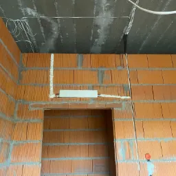 Surowy mur z cegły ceramicznej z widoczną instalacją elektryczną pod sufitem, w tym peszle i puszki, oraz fragmentem instalacji wypełnionej pianką montażową.