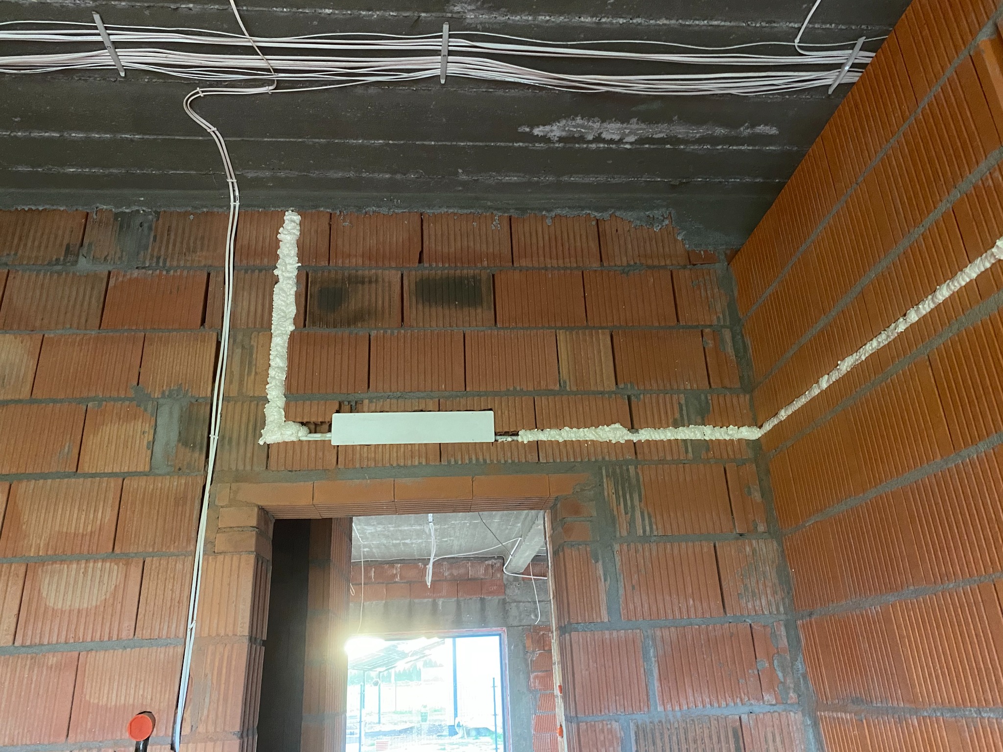 Instalacja elektryczna w budynku w trakcie budowy; widoczne przewody na suficie, puszka elektryczna zamontowana w ścianie z cegły oraz pianka montażowa uszczelniająca przejścia przewodów.