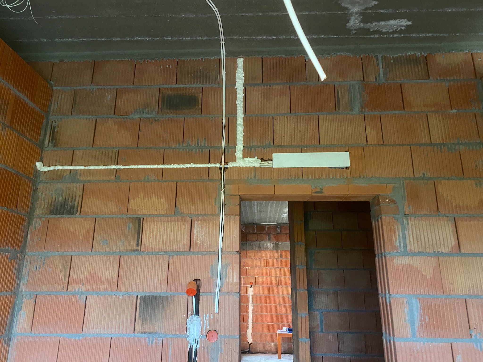 Ściana z czerwonej cegły dziurawki w trakcie instalacji elektrycznej, zabezpieczona pianką montażową, widoczne kable i puszka elektryczna, w tle widok na sąsiednie pomieszczenie.