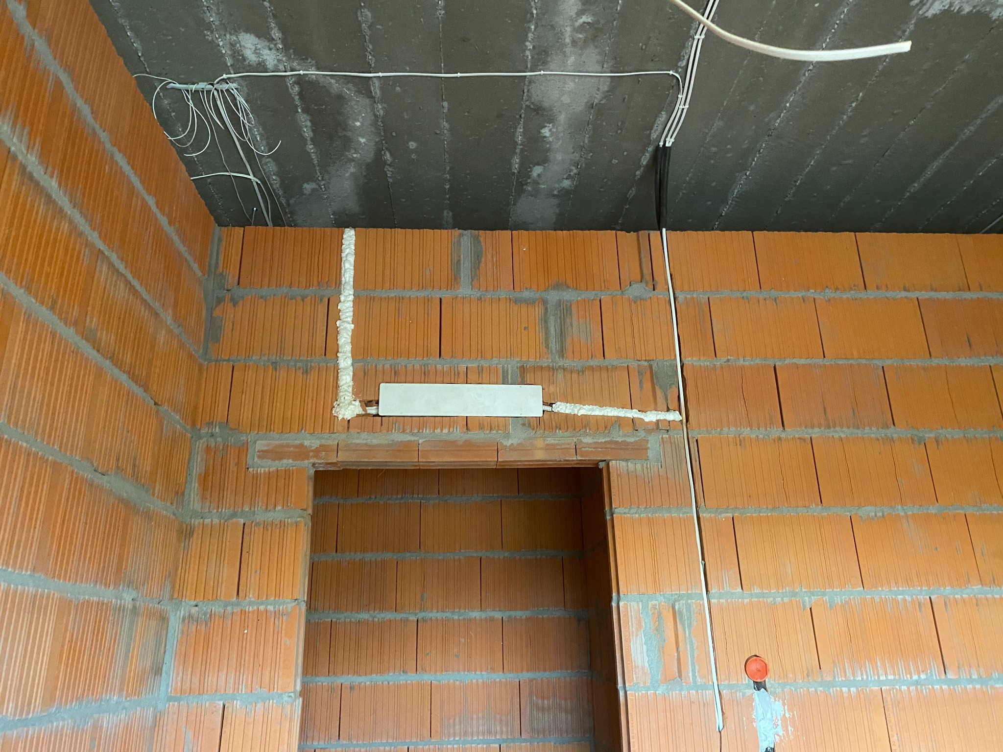 Surowy mur z cegły ceramicznej z widoczną instalacją elektryczną pod sufitem, w tym peszle i puszki, oraz fragmentem instalacji wypełnionej pianką montażową.