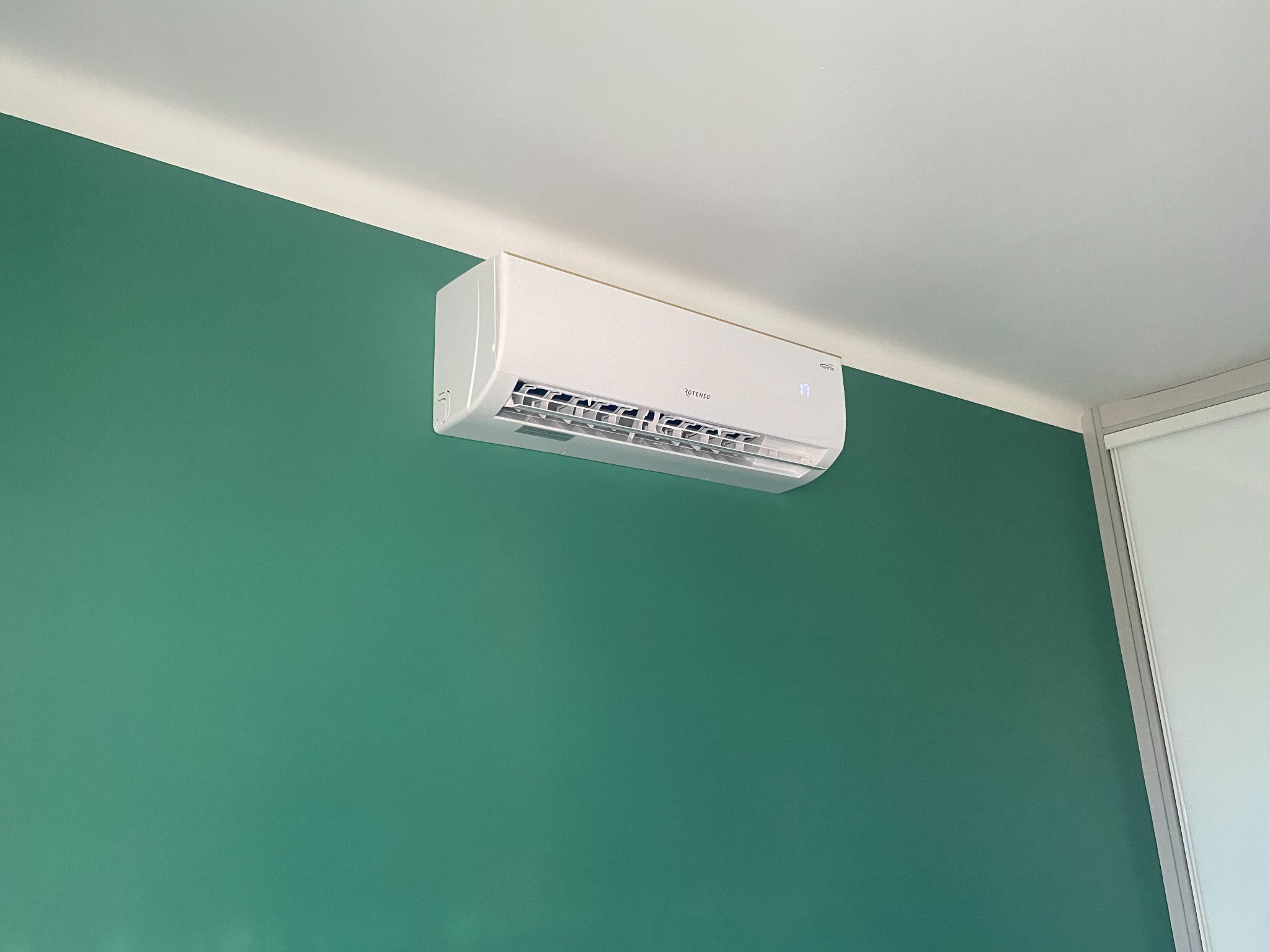 Biała klimatyzacja Rotenso zamontowana na zielonej ścianie, widoczny fragment sufitu i przesuwne drzwi w tle.