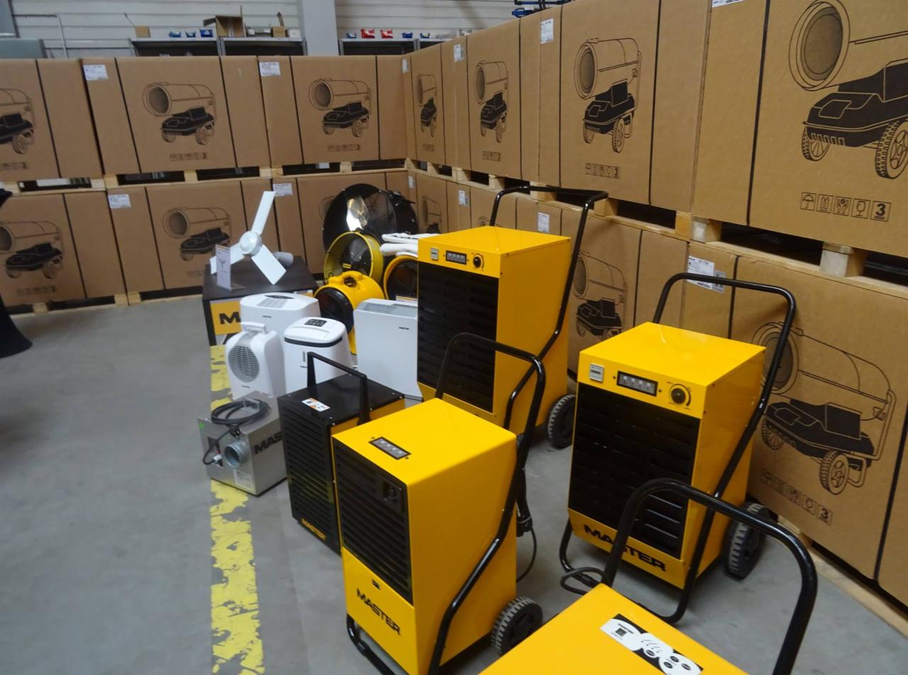 Różne modele osuszaczy i wentylatorów marki Master ustawione na tle palet z kartonami, prezentacja sprzętu w magazynie.