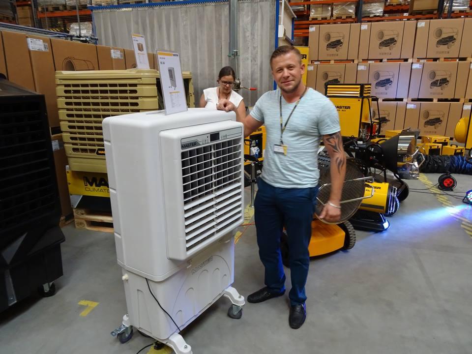 Mężczyzna z tatuażem na ramieniu prezentuje mobilny klimatyzer ewaporacyjny Master w magazynie, w tle widoczne nagrzewnice i regały z kartonami.