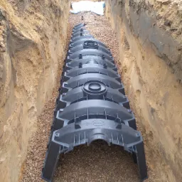 Długi, czarny tunel z tworzywa sztucznego ułożony w wykopie wypełnionym drobnym żwirem; ściany wykopu z piasku i ziemi, widoczny pojazd w tle.