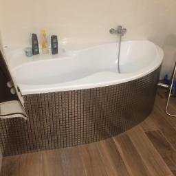 USŁUGI BUDOWLANO-REMONTOWE,LASKOWSKI PIOTR - Biała wanna obudowana mozaiką w odcieniach brązu, z widocznymi kosmetykami na krawędzi i drewnopodobną podłogą, sfotografowana z niskiego kąta.