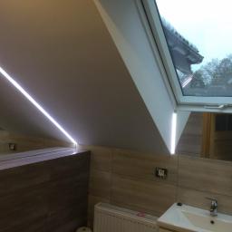 USŁUGI BUDOWLANO-REMONTOWE,LASKOWSKI PIOTR - Łazienka na poddaszu z oknem dachowym, podświetleniem LED wzdłuż skosu sufitu, płytkami w odcieniach beżu i brązu oraz białą umywalką.