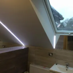 Łazienka na poddaszu z oknem dachowym, podświetleniem LED wzdłuż skosu sufitu, płytkami w odcieniach beżu i brązu oraz białą umywalką.